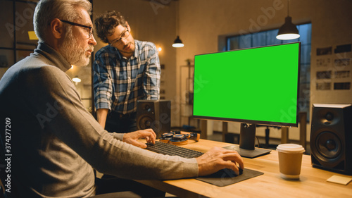 In the Evening Creative Middle Aged Man Works on a Desktop Computer Green Mock-up Screen, Has Conversation with Younger Specialist who Uses Tablet Computer. Stylish Big City Office late in the Evening