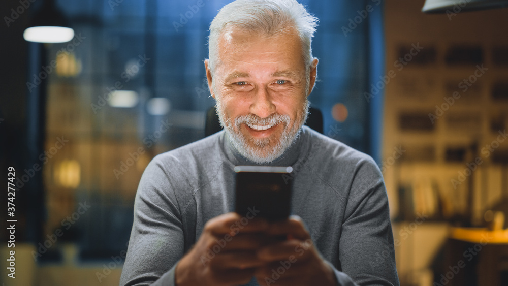 Portrait of the Handsome and Successful Middle Aged Bearded Businessman Uses Smartphone while Sitting at His Desk, Laughs and Smiles. Working from Cozy Home Office / Studio Portrait of the Handsome and Successful Middle Aged Bearded Businessman Uses Smartphone while Sitting at His Desk, Laughs and Smiles. Working from Cozy Home Office / Studio