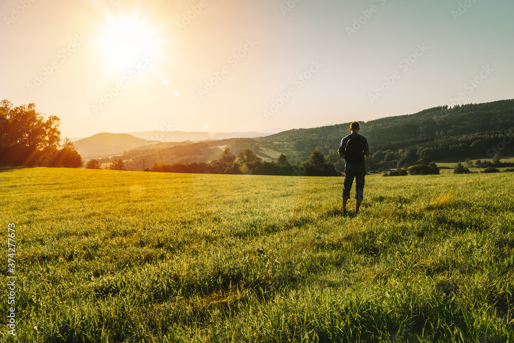 Young man with backpack walking on meadow in summer Czech landscape at sunset
