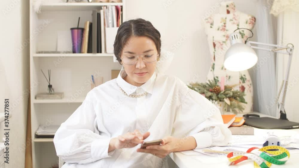 Beautiful designer woman is typing a text message on her smartphone while sitting in her home workshop. Beautiful designer woman is typing a text message on her smartphone while sitting in her home workshop.