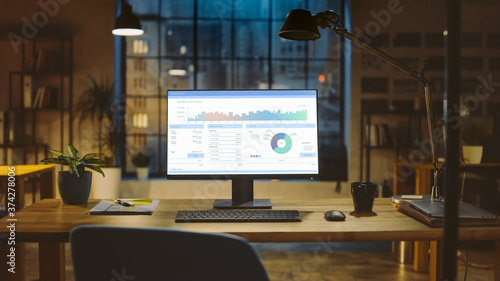 Shot of a Desktop Computer with Statistics Graph, Chart and Various Data, Showing Company Growth and Success. In the Background Warm Evening Lighting and Open Space Studio with City Window View.
