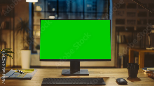 Desktop Computer with Mock-up Green Screen Standing on the Wooden Desk in the Modern Creative Office. In the Background Warm Evening Lighting and Open Space Studio with City Window View. photo