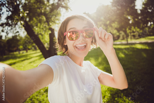 Closeup headshot photo of careless charming pretty lady shiny toothy smiling hold camera spectacles take selfie open mouth send picture friend abroad park wear sunglass white shirt outdoors