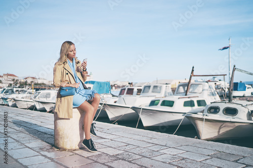 Foto Young traveling woman in coat  sitting on Split promenade sea embankment using smartphone