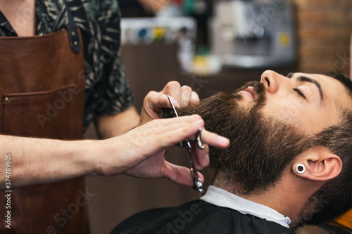 Bearded male sitting in an armchair in a barber shop while hairdresser trip his beard with scissors. Close-up of barber shearing beard to man in barbershop