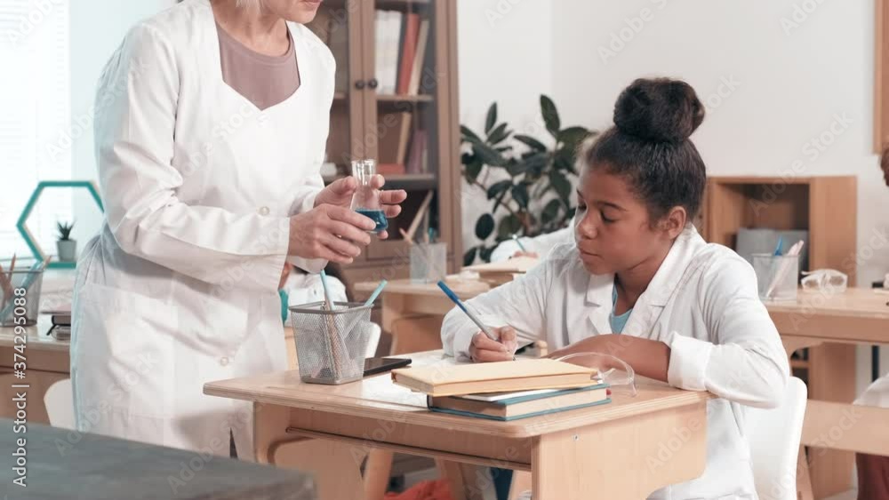 Handheld shot of woman walking up to African-American schoolgirl sitting at her desk in classroom and showing her flask with blue liquid while teaching chemistry in middle school Handheld shot of woman walking up to African-American schoolgirl sitting at her desk in classroom and showing her flask with blue liquid while teaching chemistry in middle school