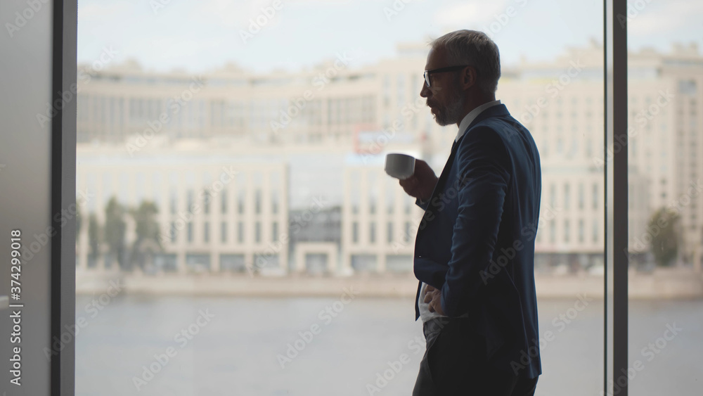 Happy older businessman taking break in office drinking coffee standing near window Happy older businessman taking break in office drinking coffee standing near window