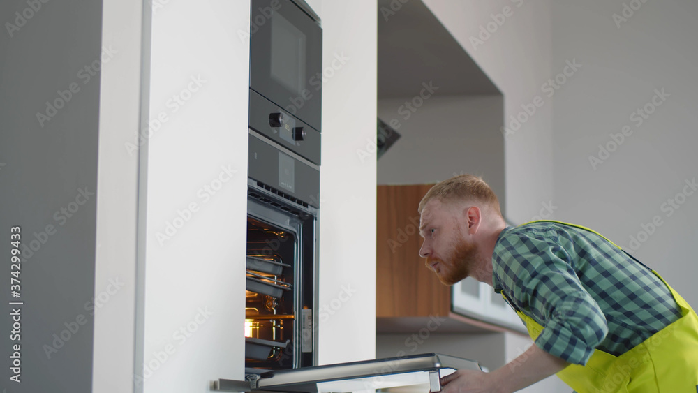 Repairman in uniform examining broken electric oven at home kitchen Repairman in uniform examining broken electric oven at home kitchen