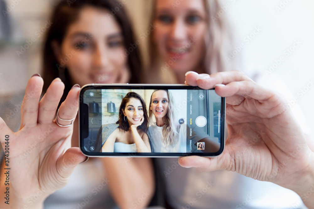 Smiling young women friends, brunette and blonde, make selfie on the phone in a cafe. Friendship and communication.