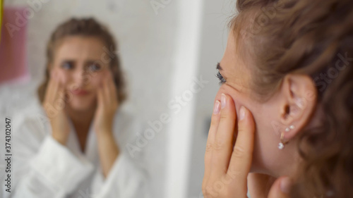 Unhappy young woman looking in mirror at home touching face