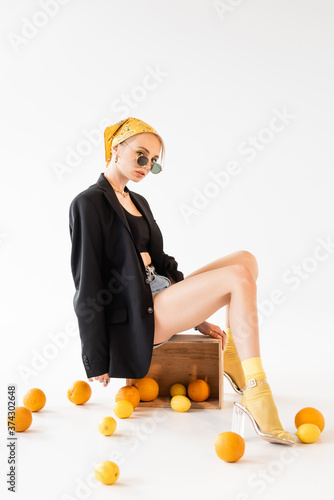 fashionable woman posing on wooden box near scattered citrus fruits on white background