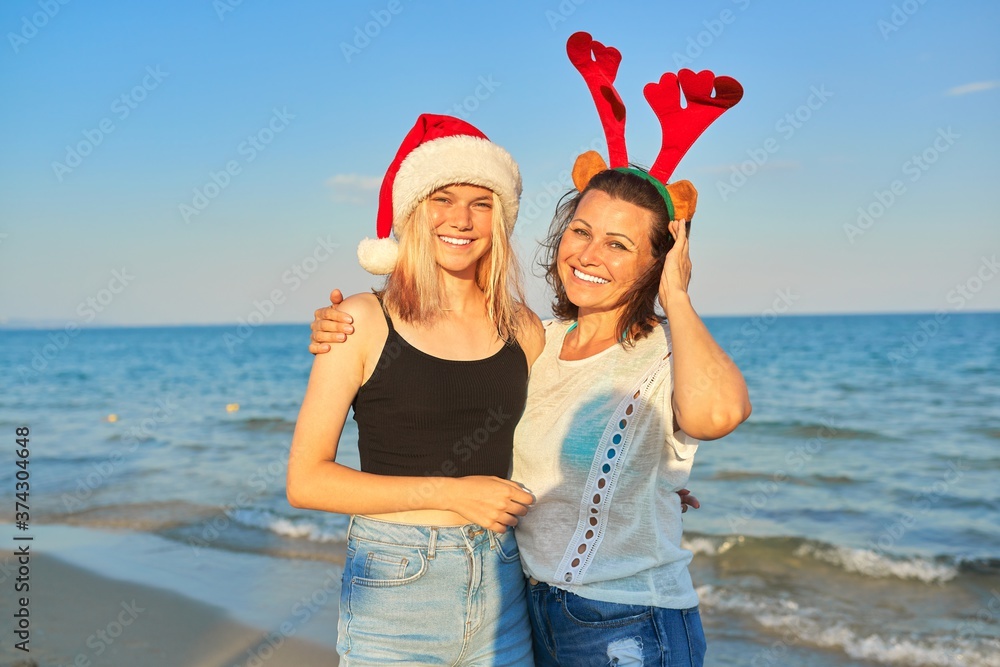 Obraz premium Happy mother and teen daughter in Santa Claus hat on beach, New Year Christmas holidays