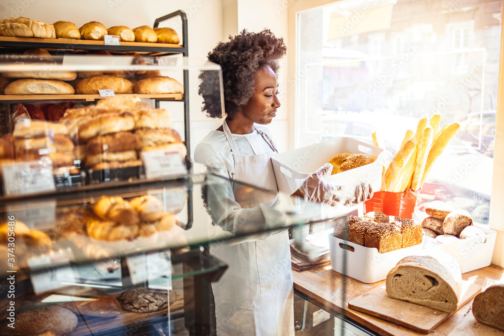 Beautiful female baker holding a tray of bread at the bakery. Young woman selling bread and pastry. Portrait of a confident young woman working in a bakery shop. Happy business owner at a bakery shop