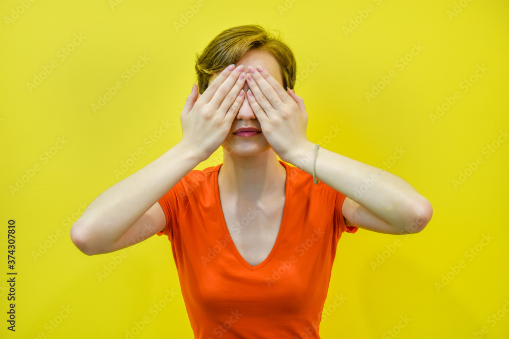 A young woman with short hair covered her eyes with her hands. Portrait of a beautiful young woman on a yellow wall background, space for text. Concept: fright, blindness, see nothing. A young woman with short hair covered her eyes with her hands. Portrait of a beautiful young woman on a yellow wall background, space for text. Concept: fright, blindness, see nothing.