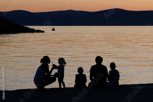 Black silhouettes of people against the background with sunset by the sea. Family is sitting on edge of the pier and relaxing. Sveti Juraj, Croatia