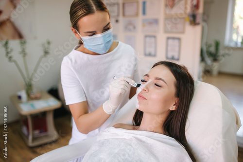Young woman cosmetologist apply white clay mask on woman s face. Beautiful brunette woman in beauty salon