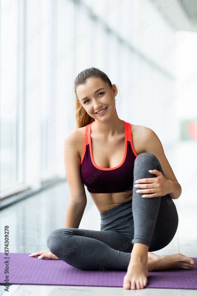 Young fit woman in activewear sitting on mat during physical exercise on background of large window in leisure center Young fit woman in activewear sitting on mat during physical exercise on background of large window in leisure center