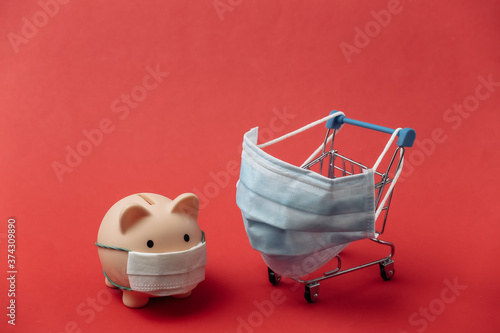 Mini supermarket trolley and piggy bank with medical mask on red background. Covid-19 pandemic