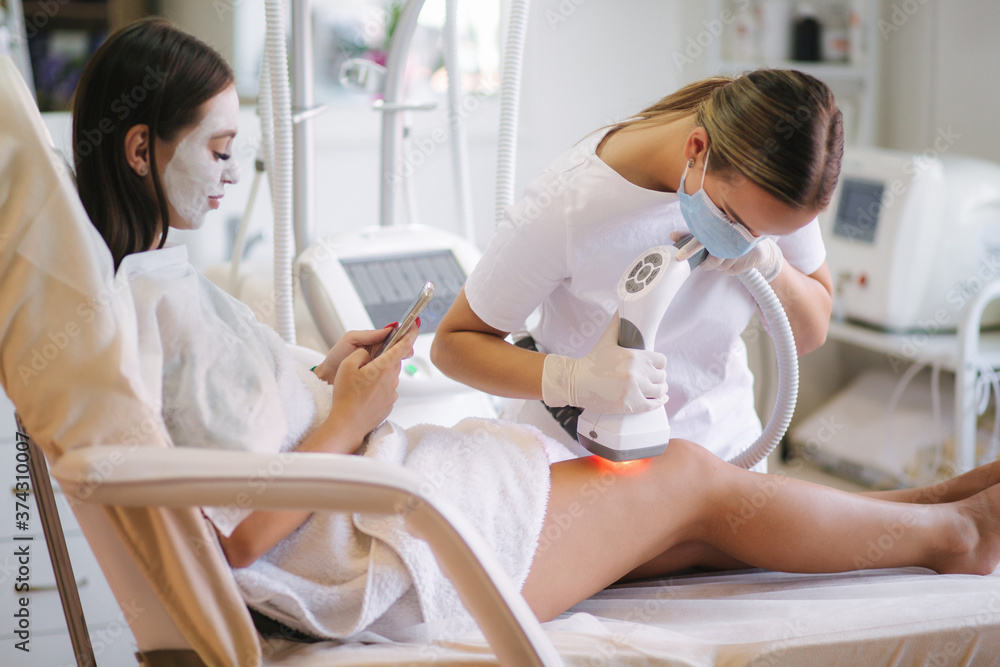 Beutiful bussines woman with white facial mask using phone while cosmetologist in white gloves performing radiofrequency lifting procedure on the stomach. Beautiful woman in medical clinic. Beautician