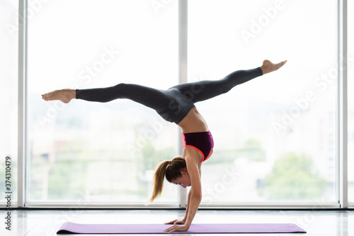 Side view of young girl gymnast doing pincha mayurasana exercise handstand pose on mat on floor of panoramic window. Concept of professional athletes gymnasts and acrobats. Advertising space