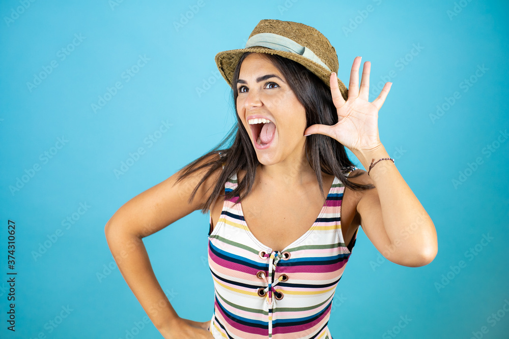 Young beautiful woman wearing swimsuit and hat over isolated blue background smiling with hand over ear listening an hearing to rumor or gossip Young beautiful woman wearing swimsuit and hat over isolated blue background smiling with hand over ear listening an hearing to rumor or gossip
