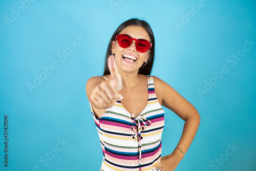 Young beautiful woman wearing swimsuit and sunglasses over isolated blue background smiling and doing the ok signal with her thumb