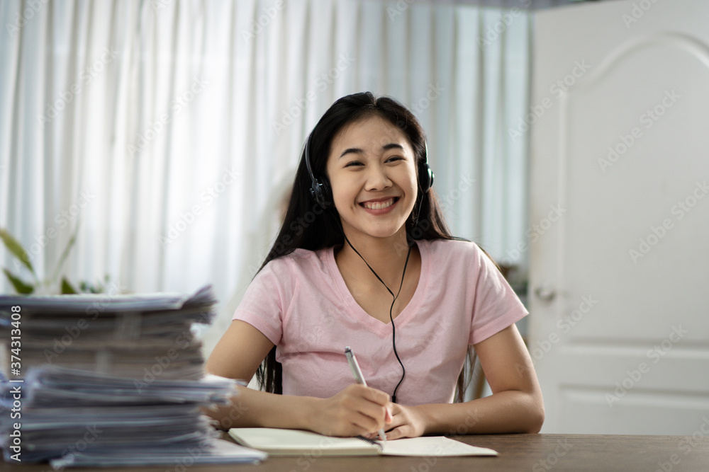 A beautiful Asian girl in pink jeans and headphones is sitting on a chair and smiling in her house writing a notebook..