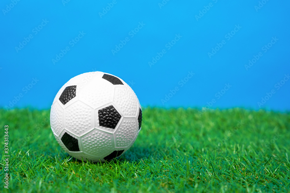 Miniature toy soccer ball lies on green grass of artificial turf of football field, blue background, front view, close up. Equipment for outdoor activities and sports.