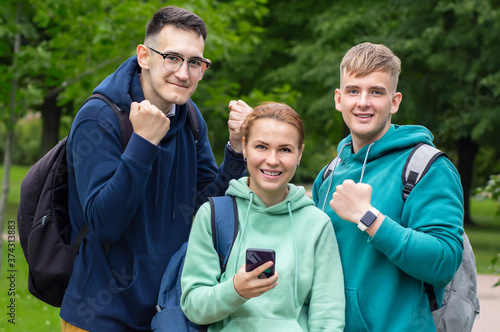 Three happy university or college students with smartphone. Friends celebrating success, the end of session, lessons. Pass exams, good results. Successful guys outdoors in park with backpacks. © Евгений Шемякин