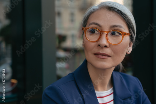 Close up portrait of pensive mature businesswoman looking away, planning start up. Beautiful middle aged woman wearing stylish eyeglasses standing outdoors, focus on face
