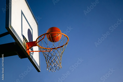 Street basketball ball falling into the hoop. Close up of orange ball above the hoop net with blue sky in the background. Concept of success, scoring points and winning. Copy space © CrispyMedia