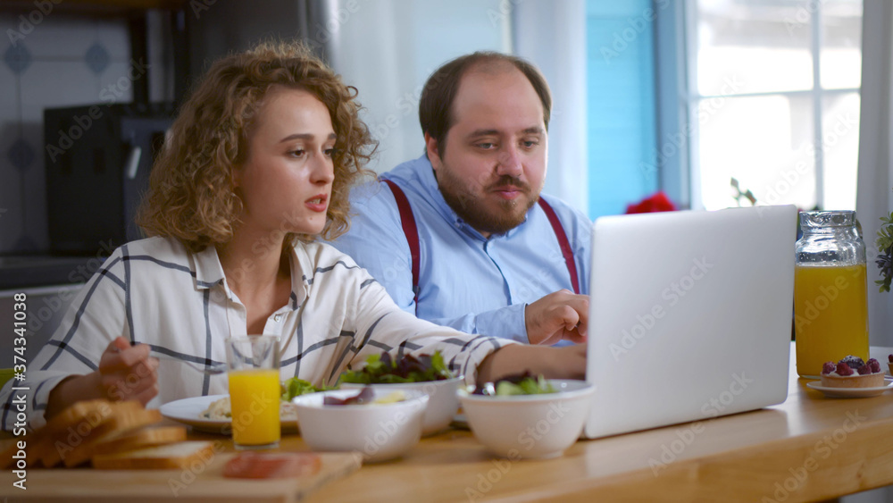 Cheerful loving couple having breakfast sitting at table in kitchen at home and looking at laptop Cheerful loving couple having breakfast sitting at table in kitchen at home and looking at laptop