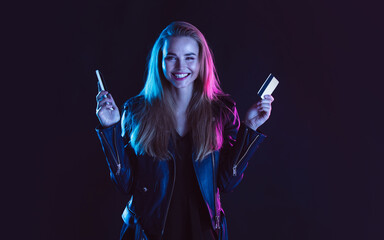 Online purchase. Portrait of young beautiful woman with card, phone in neon light on dark studio backgound. The human emotions, black friday, cyber monday, purchases, sales, finance concept. Copyspace