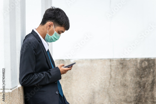 A handsome young asian business man using smartphone. Asian young attractive businessman wearing a face mask leaning against the wall.