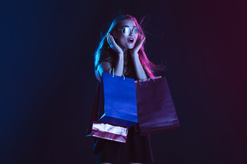 Astonished, shocked with shopping bags, cheerful. Portrait of young woman in neon light on dark backgound. The human emotions, black friday, cyber monday, purchases, sales, finance concept.