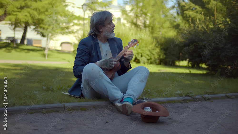Street musician beggar playing guitar asking for money or food sitting on grass in park Street musician beggar playing guitar asking for money or food sitting on grass in park
