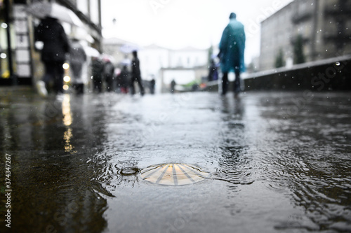 Foto Bronze scallop as a waymark in the old town of Santiago de Compostela on wet ground during rain with pilgrims in ponchos in the background