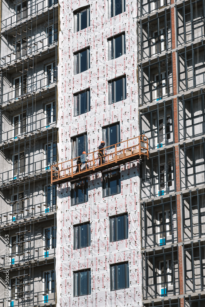 Builders on an industrial elevator are engaged in cladding and insulation of a high-rise residential building under construction. Detail of residential building under construction Builders on an industrial elevator are engaged in cladding and insulation of a high-rise residential building under construction. Detail of residential building under construction