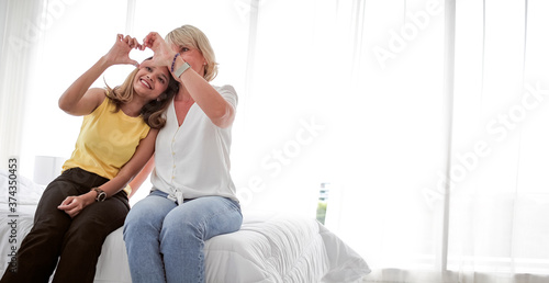 Old senior and young woman closeup show heart shape hand sign gesture, mother daughter or grandmother granddaughter in family, mature and young female sitting in white bed room
