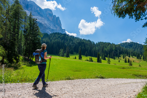 Young woman with a hat, trekking poles, a backpack hiking on a trail through green meadows in the Italian Alps. Dolomite peaks are visible in the background. Val Badia, South Tyrol - Italy