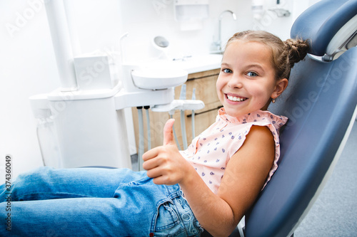 Happy little girl with a perfect white smile showing thumbs up  she sitting in a dental chair. She have healthy teeth