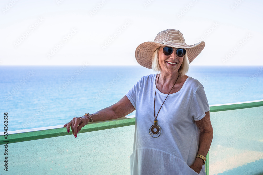 Senior woman leaning on a terrace railing looking out to sea wearing a straw hat and white dress