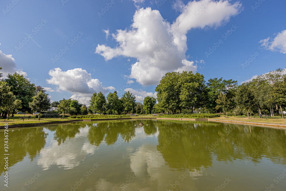 Obraz premium Pond with a mirror reflection in the water surrounded by lush trees in the city park, sunny summer day with a blue sky and white clouds in Sittard, South Limburg, Netherlands
