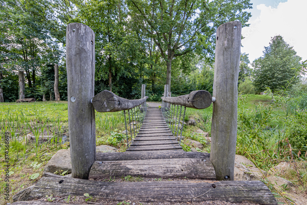 Small suspension bridge with huge wooden logs over the Keutelbeek stream with lush trees in the background, sunny summer day with a blue sky and white clouds in Sittard in South Limburg, Netherlands