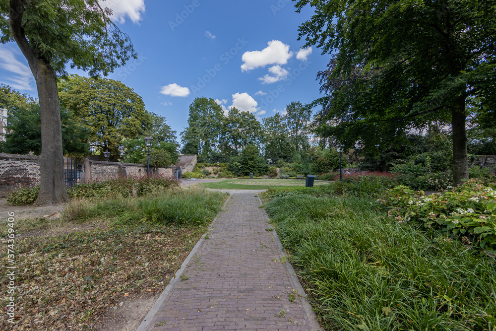 Cobbled pedestrian sidewalk between green grass in the Isabelle garden (Jardin d'Isabelle) with lush trees in the background, sunny summer day with a blue sky in Sittard, South Limburg, Netherlands