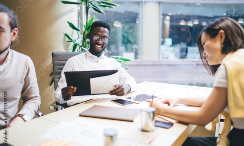 Smiling dark skinned male and female diverse group sitting together on meeting table working on strategy and planning creation, prosperous crew of employees satisfied with cooperation on brainstorming