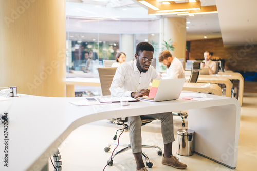 Concentrated dark skinned man typing on laptop computer making online research on web pages, 20s serious young male manager making planning and sending mails sitting at working table in office