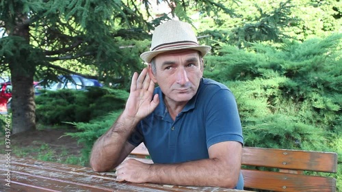 Middle Age Man Sitting o Bench in Park and Holding Left Hand Behind Ear Indicating That He Can Not Hear Well