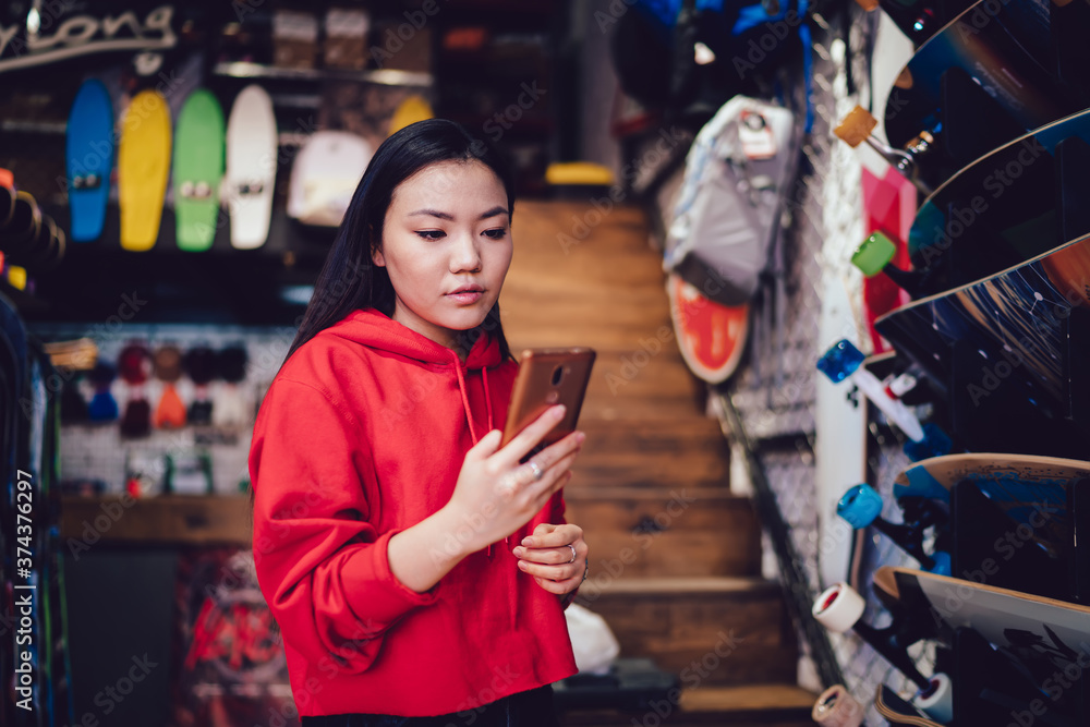 Millennial female teenager using cellphone for browsing web store and compare prices during shopping day for choosing youthful skateboard, Chinese generation Z making banking payment for purchase