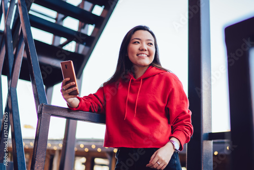 Cheerful female with modern smartphone gadget standing at urban setting and smiling, funny millennial woman dressed in stylish teenage clothing looking away during cellular phoning at leisure